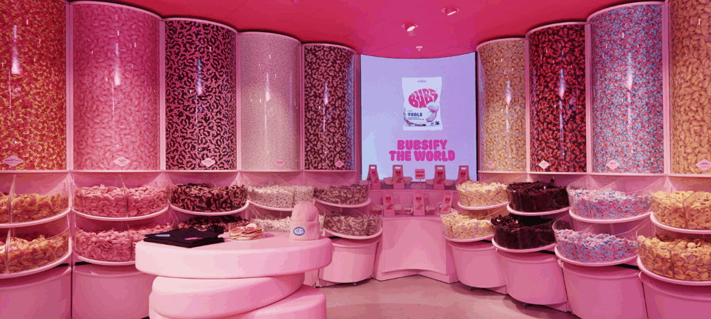 Färgglatt rosa godisbutiksinrum med höga genomskinliga behållare fyllda av godis längs väggarna och en skärm med texten "Bubsify the world".